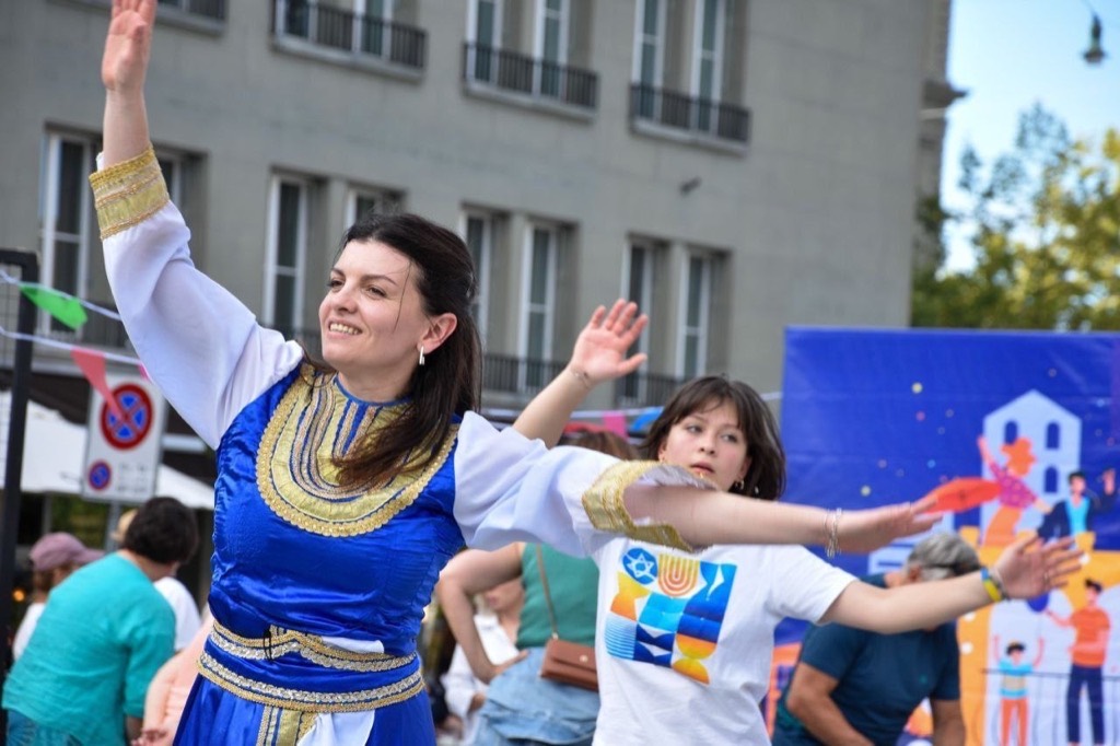 Woman in a blue embroidered dress dancing at an outdoor Jewish cultural festival, with a child in a branded shirt nearby