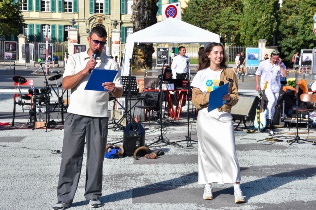 Two presenters with microphones and clipboards on stage with musical equipment in a city square